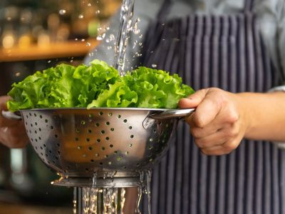 Person Cleaning Leafy Greens In A Metal Colander