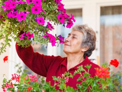 Older Lady Smelling Flowers From A Hanging Basket