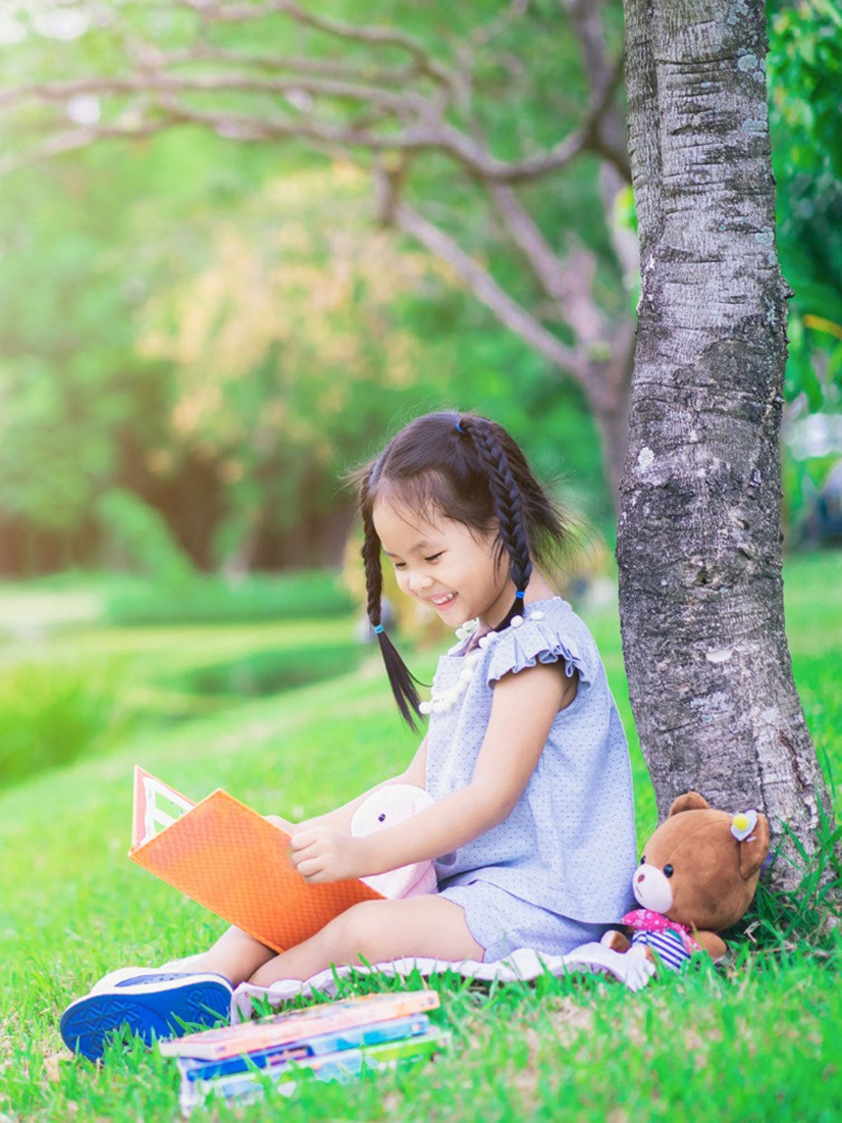 A smiling girl with stuffed animals reads a book under a tree