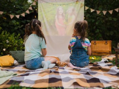 Two girls sit outside on a blanket watching a movie projected on a hanging sheet