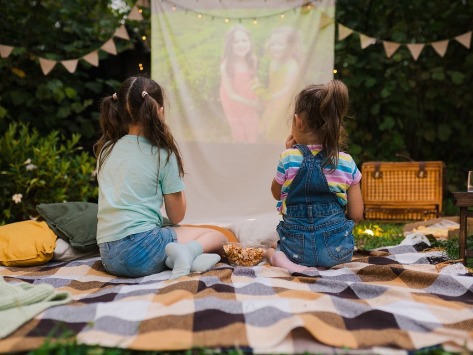 Two girls sit outside on a blanket watching a movie projected on a hanging sheet