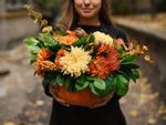 A woman holds an arrangement of green and orange fall flowers in a pumpkin