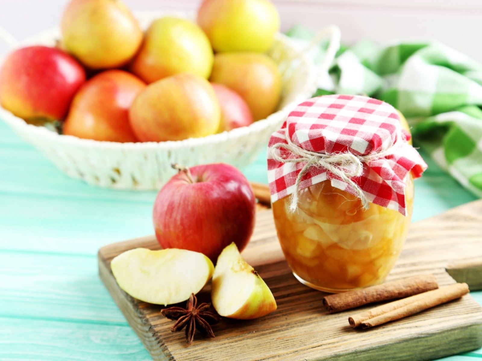 Sliced apple and cinnamon sticks next to a jar of preserved apples, with a bowl of whole apples in the background