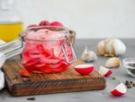 A jar of pickled radishes sits on a cutting board next to sliced radishes, peppercorns, and garlic cloves