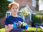 A red haired boy standing outside a house smiles and holds a blue plastic bottle with pansies growing in it