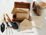 A small box of plain paper envelopes with the names of herbs printed on them, next to small planting pots and gardening tools