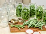 Many glass jars, green beans, and canning lids sit on a table with a cutting board