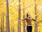 A woman stands with her eyes closed and arms outstretched in a forest of trees with yellow leaves