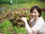A smiling woman in a garden holds up a large bunch of red lettuce