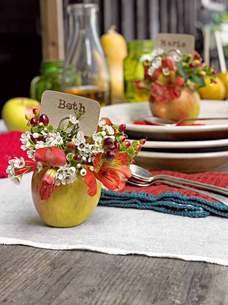 A table laid with plates and cutlery, and apples hollowed out for use as vases filled with autumn flowers and berries, and place names